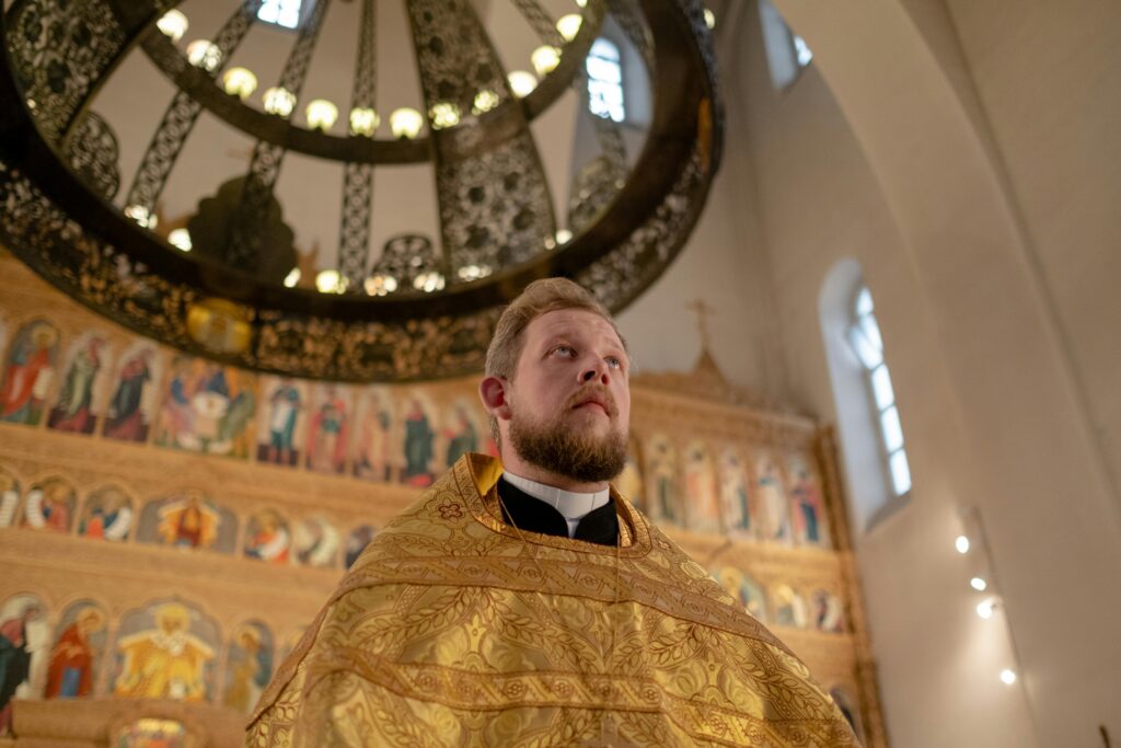 Low-angle view of an Orthodox priest in a richly decorated church with an iconostasis and ornate ceiling.